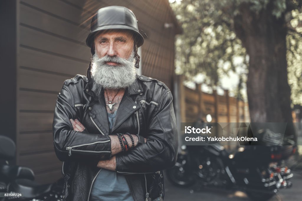 Cheerful smiling old man in helmet Glad aged bearded biker wearing leather jacket is standing near motorbikes. He crossing hands and looking at camera with smile. Portrait. Copy space Biker Stock Photo