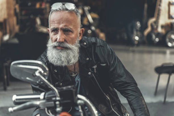Tired glance of old man on bike Attentive aged bearded biker is sitting on motorbike and looking aside with seriousness. He locating near open garage. Portrait biker stock pictures, royalty-free photos & images
