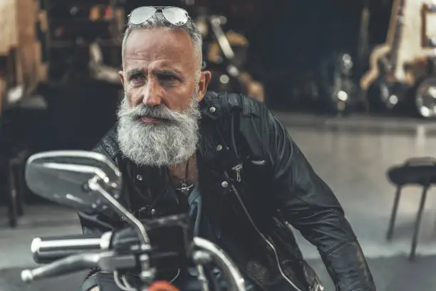 Attentive aged bearded biker is sitting on motorbike and looking aside with seriousness. He locating near open garage. Portrait