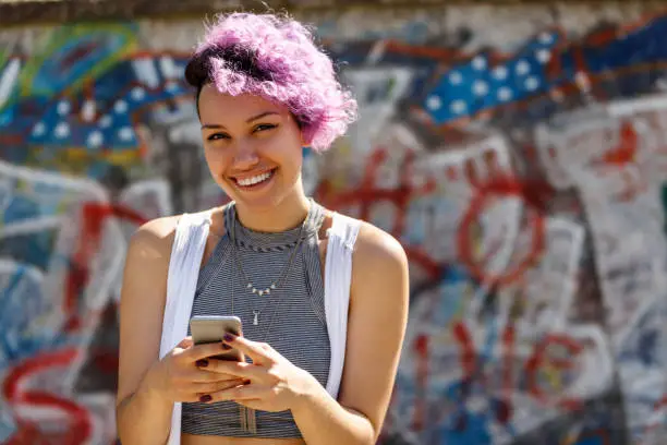 Photo of Smiling young hipster girl using smart phone against graffiti wall