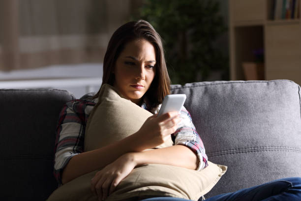 Sad girl reading phone message at home in the dark Sad girl holding pillow and reading phone message in the dark sitting on a couch at home waiting telephone on the phone frustration stock pictures, royalty-free photos & images