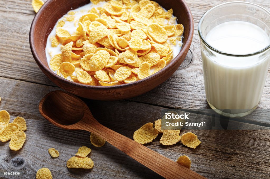 Cornflakes on a worn wooden background Cornflakes in a brown clay plate on a worn wooden background, next is a glass of milk and a spoon, and several flakes are scattered Corn Flakes Stock Photo