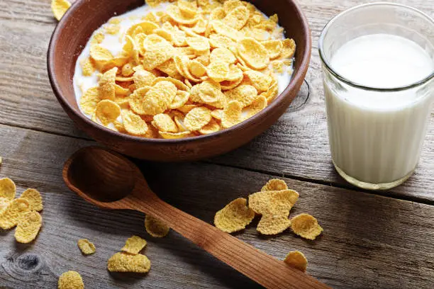 Cornflakes in a brown clay plate on a worn wooden background, next is a glass of milk and a spoon, and several flakes are scattered