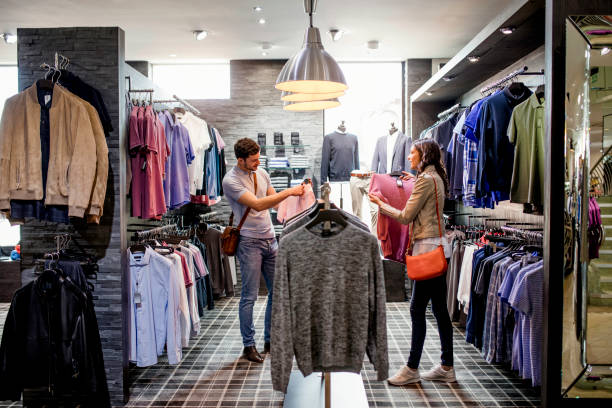He is After a New Pink Shirt Young couple standing in clothes store with rails of shirts hanging around them. The man is holding a new pink shirt while the woman shows him another clothing store stock pictures, royalty-free photos & images