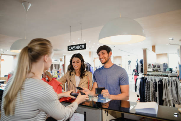 Paying For Her New Dress A young man in a fashionable clothing store at the checkout making a payment. A woman at his side smiles while placing a dress on the counter clothing store credit card stock pictures, royalty-free photos & images