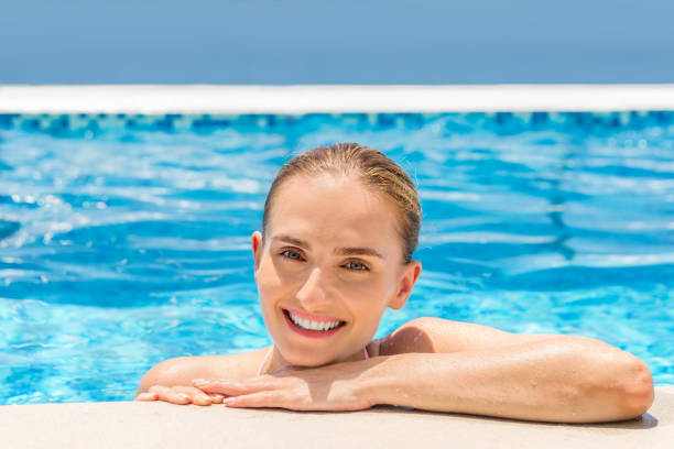 Cute young woman in luxury pool Horizontal color portrait of young woman resting at the edge of infinity pool and smiling at camera. Serenity, wellbeing and vacation. women exercising swimming pool young women stock pictures, royalty-free photos & images