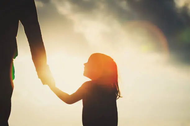 Photo of silhouette of little girl holding parent hand at sunset
