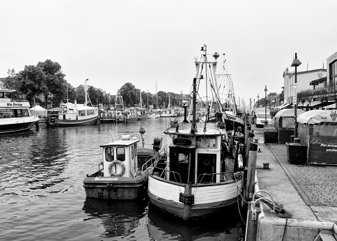 Fishing boats and leisure craft moored each side of the Alter Strom canal in Warnemünde. Only a few people are visible on a damp and unpleasant day. (Black and white image with added grain.)