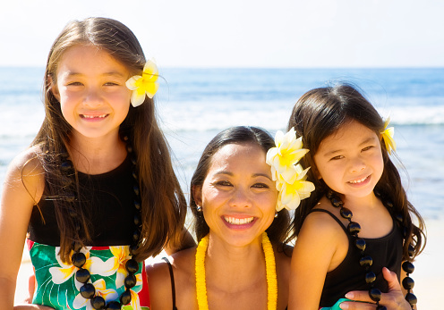 Portrait of a Hawaiian Polynesian family outdoor in the tropical beach. Two young girls with their mother posing, smiling and looking at camera half body portrait.