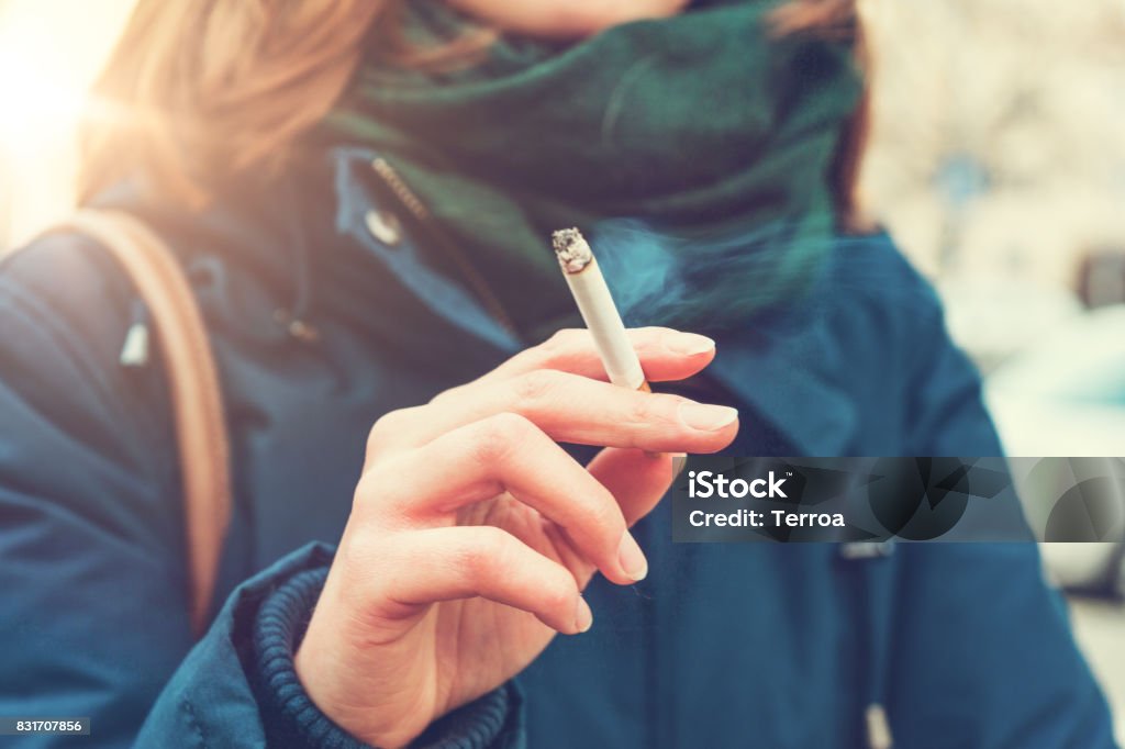 Young woman enjoying a cigarette Young woman enjoying a cigarette outdoors holding it between her fingers, low angle view against the chest of a warm autumn jacket in a smoking and tobacco concept Smoking Issues Stock Photo