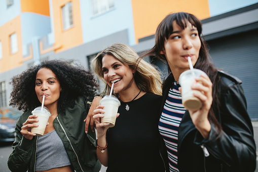 Outdoors shot of three young females having ice coffee on city street. Multiracial group of women friends drinking coffee.
