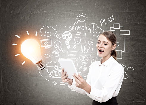 Happy blond businesswoman looking at her tablet screen and screaming with joy. Blackboard with a shining light bulb and a business scheme drawn on it.