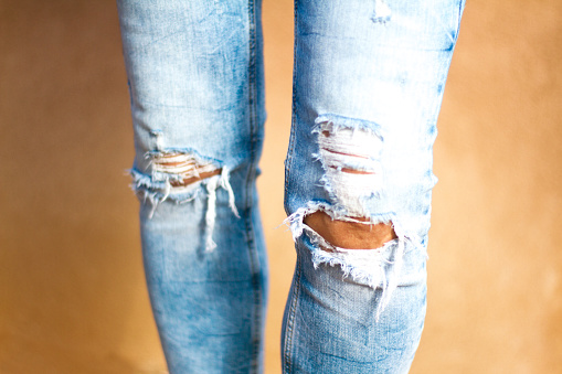 Close-up of a woman’s knees in fashionably ripped/distressed/shredded blue jeans, with yellowish textured wall background. Copy space available.