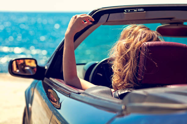 istock Beautiful blond smiling young woman in convertible top automobile looking sideways while parked near ocean waterfront 831836784