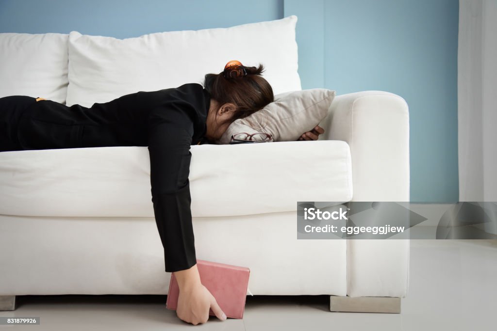 Tired Asian Business woman. Exhausted, Tired Asian Business woman in black shirt holding notebook and sleeping on white sofa with blue wall. Stress from overtime working concept. Women Stock Photo