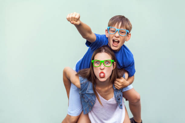 Super man style! The freckled brother climbed up on the back of a older cute sister. Making funny crazy face, tongue out and looking at camera. Crazy emotions and feelings. Super man style! The freckled brother climbed up on the back of a older cute sister. Making funny crazy face, tongue out and looking at camera. Indoor studio shot blue and green braces stock pictures, royalty-free photos & images