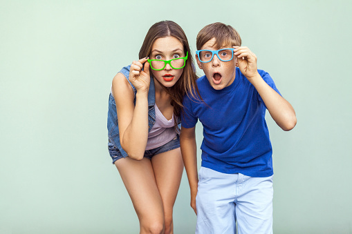 Eyewear concept. WOW faces. Young sister and brother with freckles on their faces, wearing trendy glasses, posing over light green background together. Looking at camera with surprised face. Studio shot