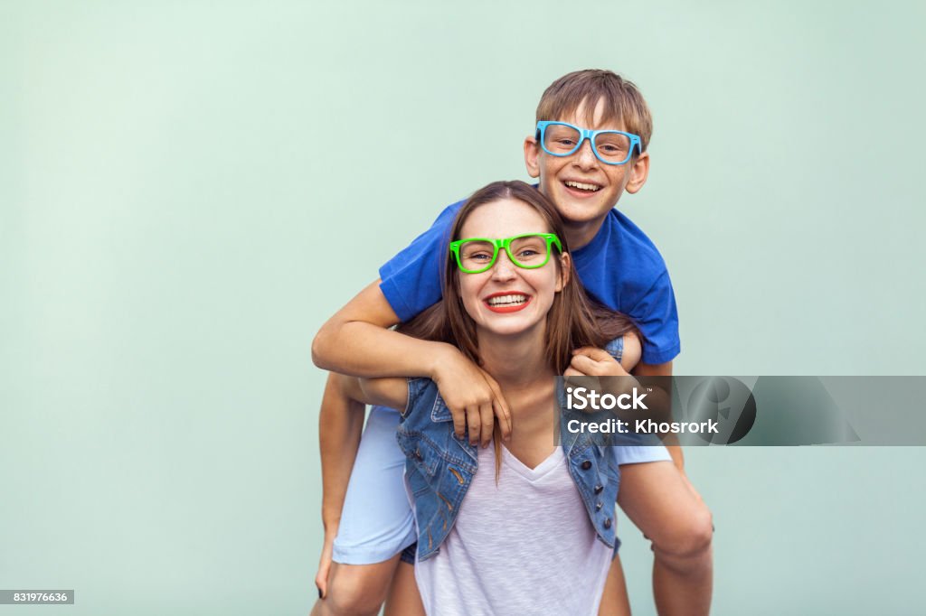 The gorgeous freckled brother and sister in casual t shirts wearing trendy glasses and posing over light blue background together. Brother climbed up on the back of a cute sister. Hipster style. Eyewear concept. The gorgeous freckled brother and sister in casual t shirts wearing trendy glasses and posing over light blue background together. Brother climbed up on the back of a cute sister. Studio shot Brother Stock Photo