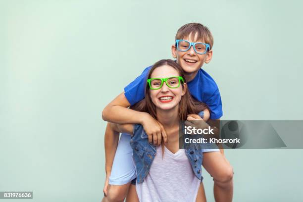 The Gorgeous Freckled Brother And Sister In Casual T Shirts Wearing Trendy Glasses And Posing Over Light Blue Background Together Brother Climbed Up On The Back Of A Cute Sister Stock Photo - Download Image Now