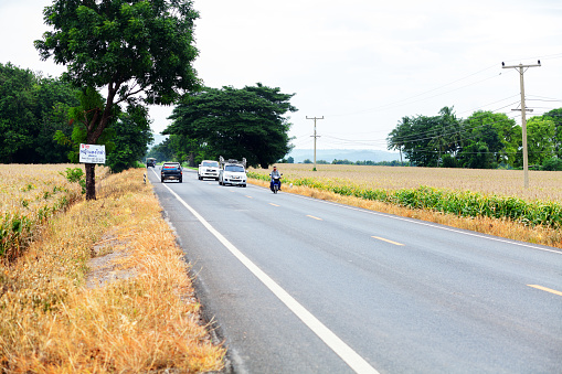 Some traffic on road in Saraburi province, road is passing landscape with corn fields. There are some cars and a thai man on motorcycle. Scene is in south east of Sap Sanun.