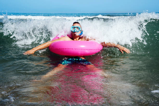 a young caucasian man wearing a diving mask, a snorkel and a pink swim ring into the seawater, next to the seashore