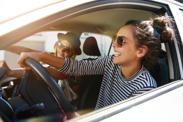 Two laughing young girlfriends driving together in a car Two attractive young girlfriends wearing sunglasses talking and laughing together while driving in a car through the city on a sunny day driving stock pictures, royalty-free photos & images