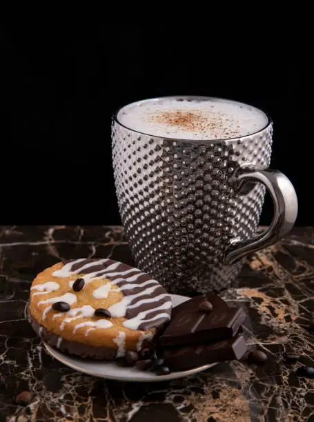 Large pimpled silver cup of coffee and cakes bisquits chocolate and coffee beans on the granite reflective surface and black background
