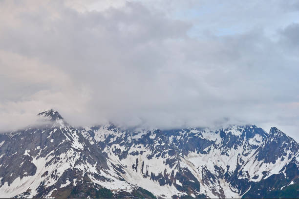 Brilliant midday sun shines in a clear blue sky above a panoramic view of the snow-covered Alpine mountain peaks. In the foreground, patches of snow and brown vegetation cover a mountain ridge. A thin airplane contrail is visible in the upper right corner of the sky. The mountain range stretches across the horizon with multiple layers of peaks fading into the distance.