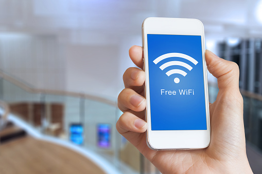 Close-up of hand holding smartphone with free WiFi hotspot icon on the screen to connect to wireless internet, public building interior in background