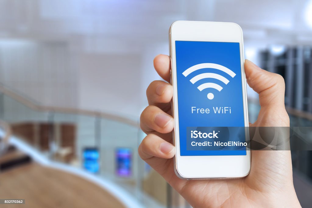 Closeup hand holding smartphone with free WiFi hotspot on screen Close-up of hand holding smartphone with free WiFi hotspot icon on the screen to connect to wireless internet, public building interior in background Wireless Technology Stock Photo