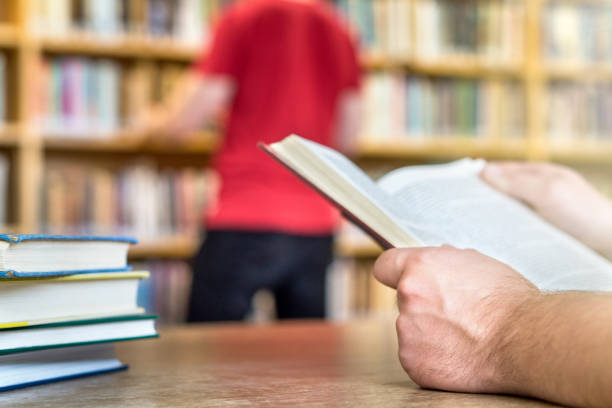 gente en la biblioteca pública o escolar en el colegio o universidad. búsqueda y lectura. concepto de servicio de educación, el estudio y la literatura. manos sosteniendo el libro con estantería y cliente en el fondo - baccalaureate fotografías e imágenes de stock