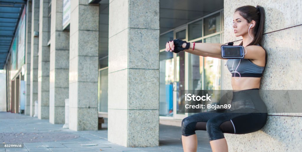 Young sporty girl doing wall sitting exercise urban outdoors. Surrounding Wall Stock Photo