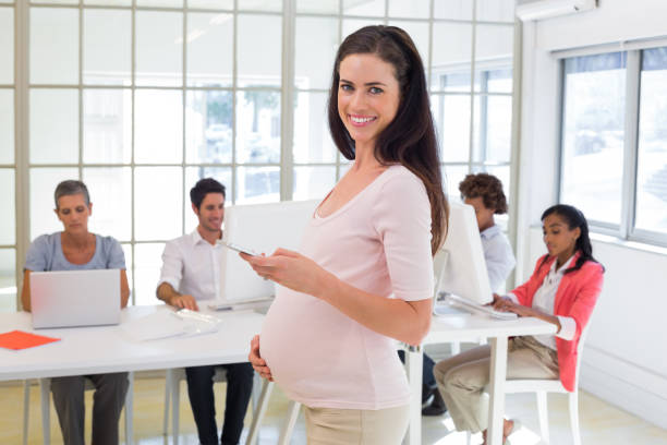 istock Pregnant office worker touches bump and texts on phone and smiles at camera 832259042