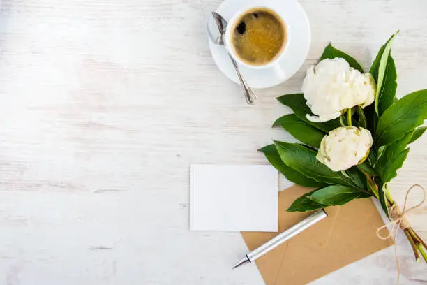 Photo of Top view of an empty greeting card, kraft envelope, peonies bouquet and a cup of coffee over white wooden rustic background.