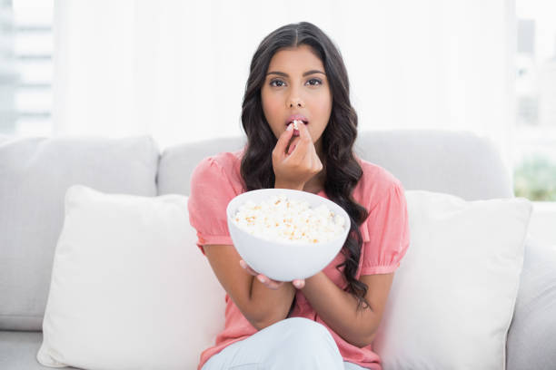 istock Frowning cute brunette sitting on couch holding popcorn bowl 832682656