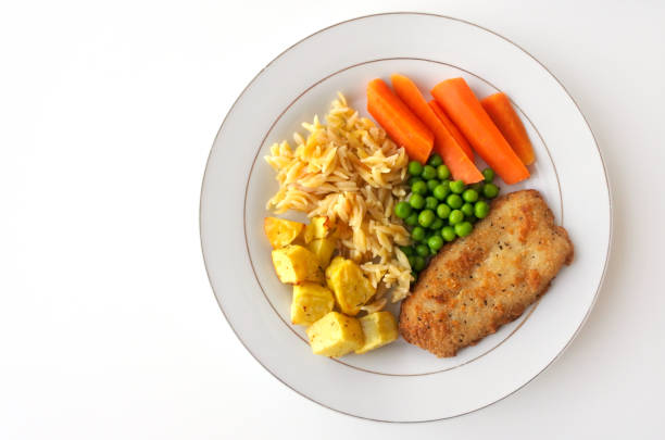 Flat lay view of lemon pepper crumbed Hoki fish fillet served with roasted potatoes, pees, carrots and orzo on a white plate. Food background and texture. Copy space