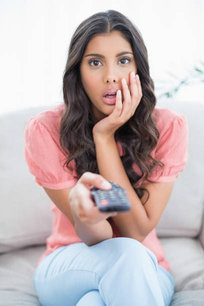 istock Shocked cute brunette sitting on couch holding remote 832794150