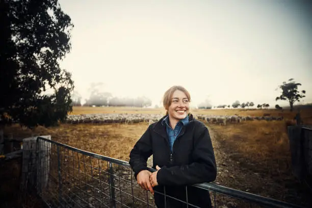 Shot of a young woman standing at a farm gate with a flock of sheep in the background
