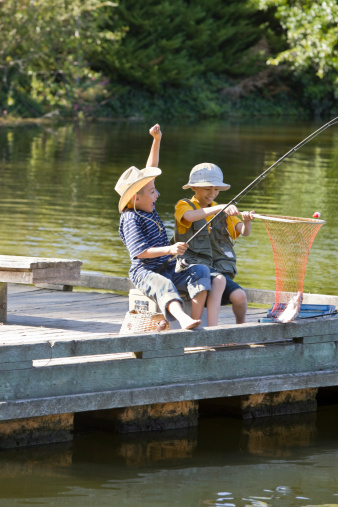 Two boys excitedly pull fish out of lake with fishing pole and net