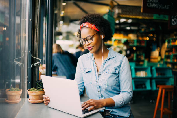 Focused young African woman working online in a cafe Focused young African woman sitting alone at a counter in a cafe working on a laptop and listening to music on earphones coffee shop stock pictures, royalty-free photos & images