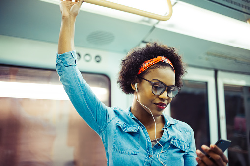 Smiling young African woman listening to music on her cellphone while standing on a subway train during her daily commute