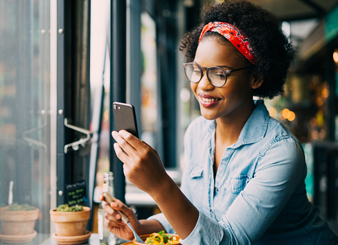 Attractive young African woman smiling and reading texts on a cellphone while sitting alone at a counter in a cafe enjoying a meal