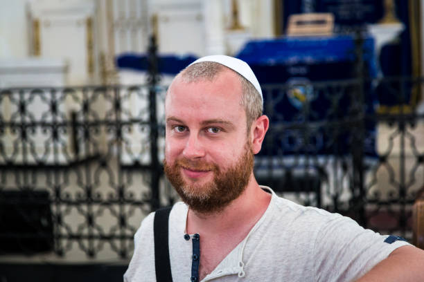 Young Jewish man wearing skull cap inside synagogue Close up image depicting a young caucasian Jewish adult man in his late 20s or early 30s inside a synagogue. He is looking at the camera and smiling, and he is wearing the traditional Jewish skull cap - otherwise known as a kippah or yarmulke - on his head. The man has a beard and the background of the synagogue is blurred out of focus. Horizontal colour image with copy space. rabbi photos stock pictures, royalty-free photos & images