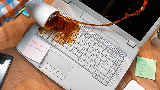 Cup of coffee spilling on white laptop in office desk.