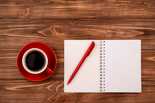 A red cup of coffee on the table. Blank notebook and a red pen on a wooden background, top view.