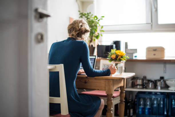 femme est assise dans une cuisine et travaille sur un ordinateur portable de la maison - rear view women back back of head photos et images de collection