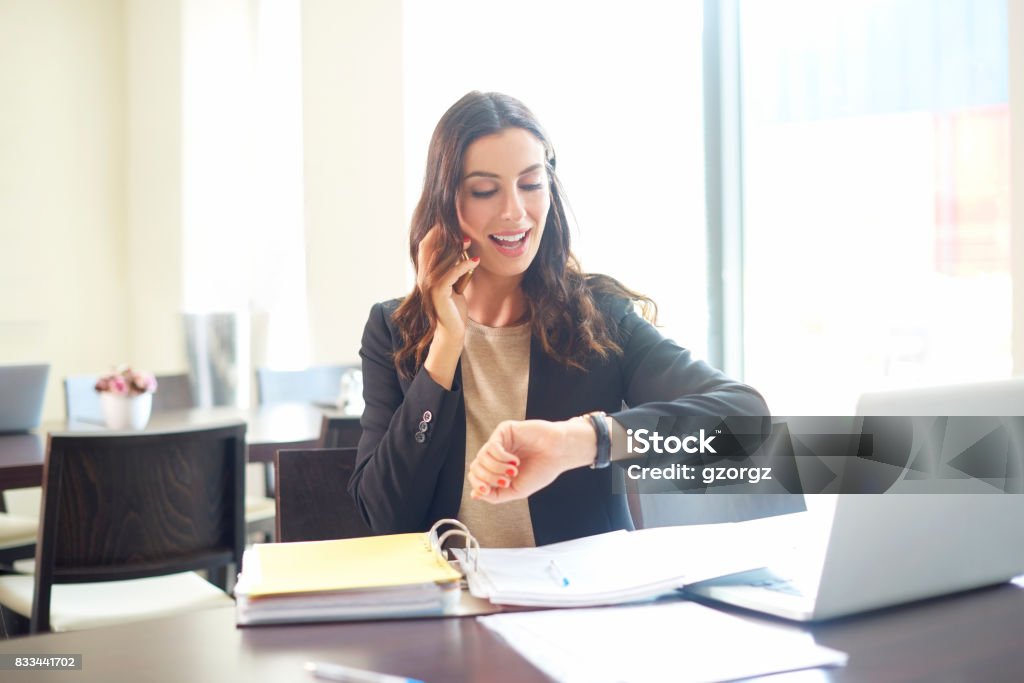 Making an appointment Shot of a young businesswoman sitting at office desk in front of laptop while using cellphone and checking time. Time Stock Photo