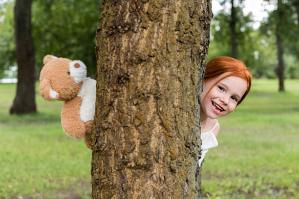 happy redhead girl holding teddy bear and hiding behind tree in park