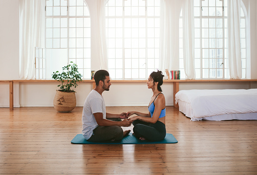 Side view of beautiful young couple doing yoga at home. They are sitting together on the yoga mat holding hands.