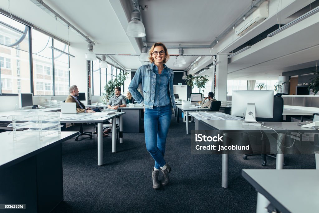 Young businesswoman standing in office with colleagues Young businesswoman standing in office with colleagues discussing in background. Happy female executive standing in workplace. Full Length Stock Photo